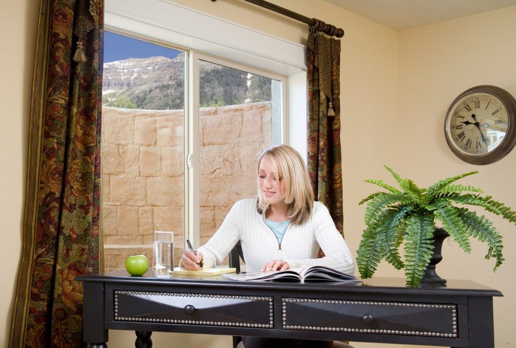 Woman studying at a desk with an egress window, enhancing natural light and safety in a below-ground space, surrounded by a plant and glass of water.