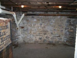 Basement interior with exposed stone walls, overhead beams, and plumbing pipes, illustrating a space in need of waterproofing and dehumidification solutions.