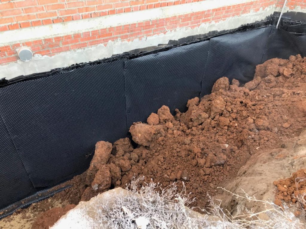 Excavated area showing protective waterproof membrane installed around foundation for exterior basement waterproofing, with exposed soil and brick wall in background.