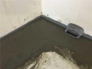 Concrete floor with a newly applied layer in a basement corner, featuring a drainage system component, illustrating interior waterproofing methods to prevent water damage.