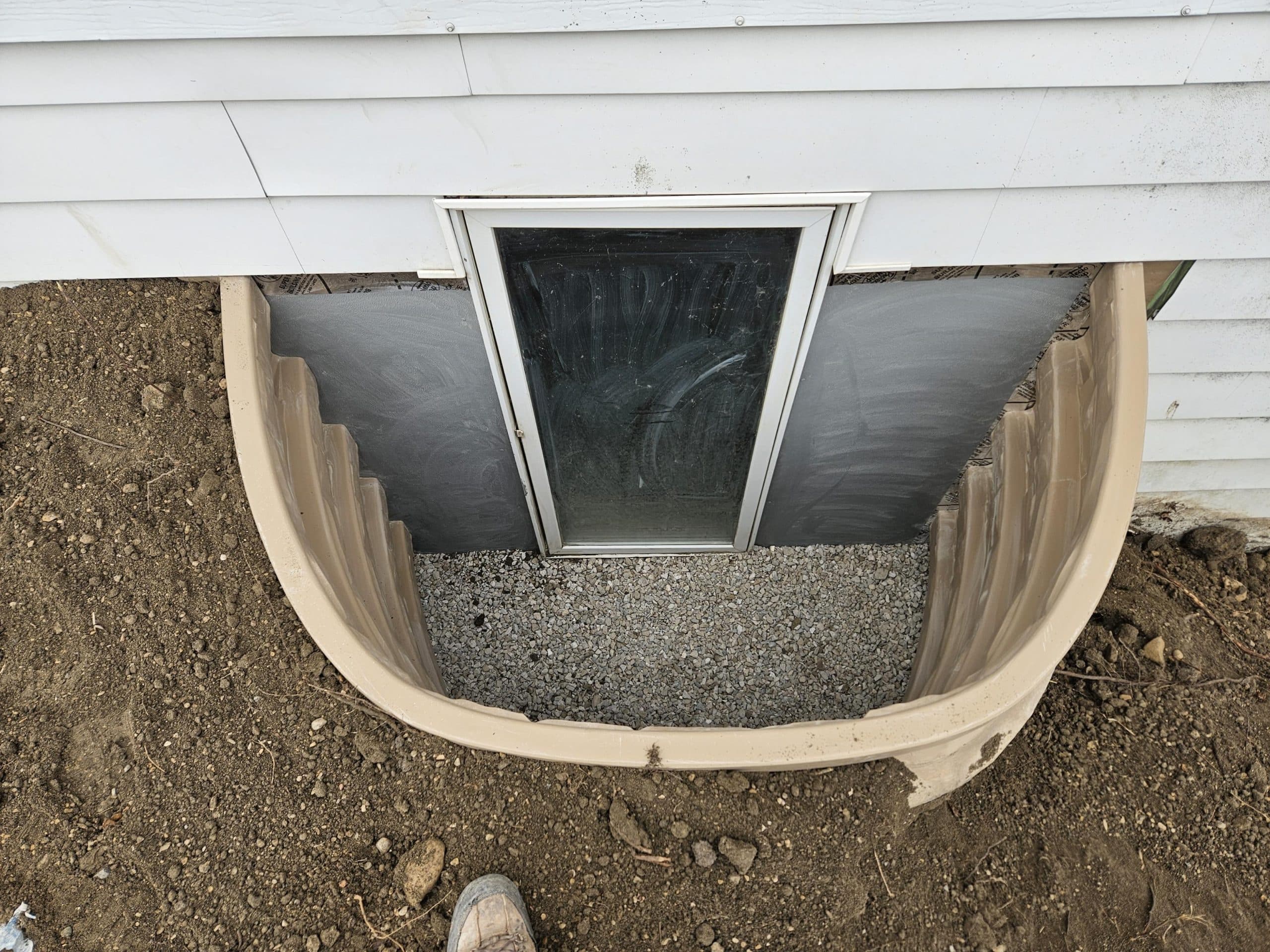 Egress window installation with a beige well and gravel base, showcasing the new window and surrounding dirt, emphasizing safety and compliance in basement renovations.