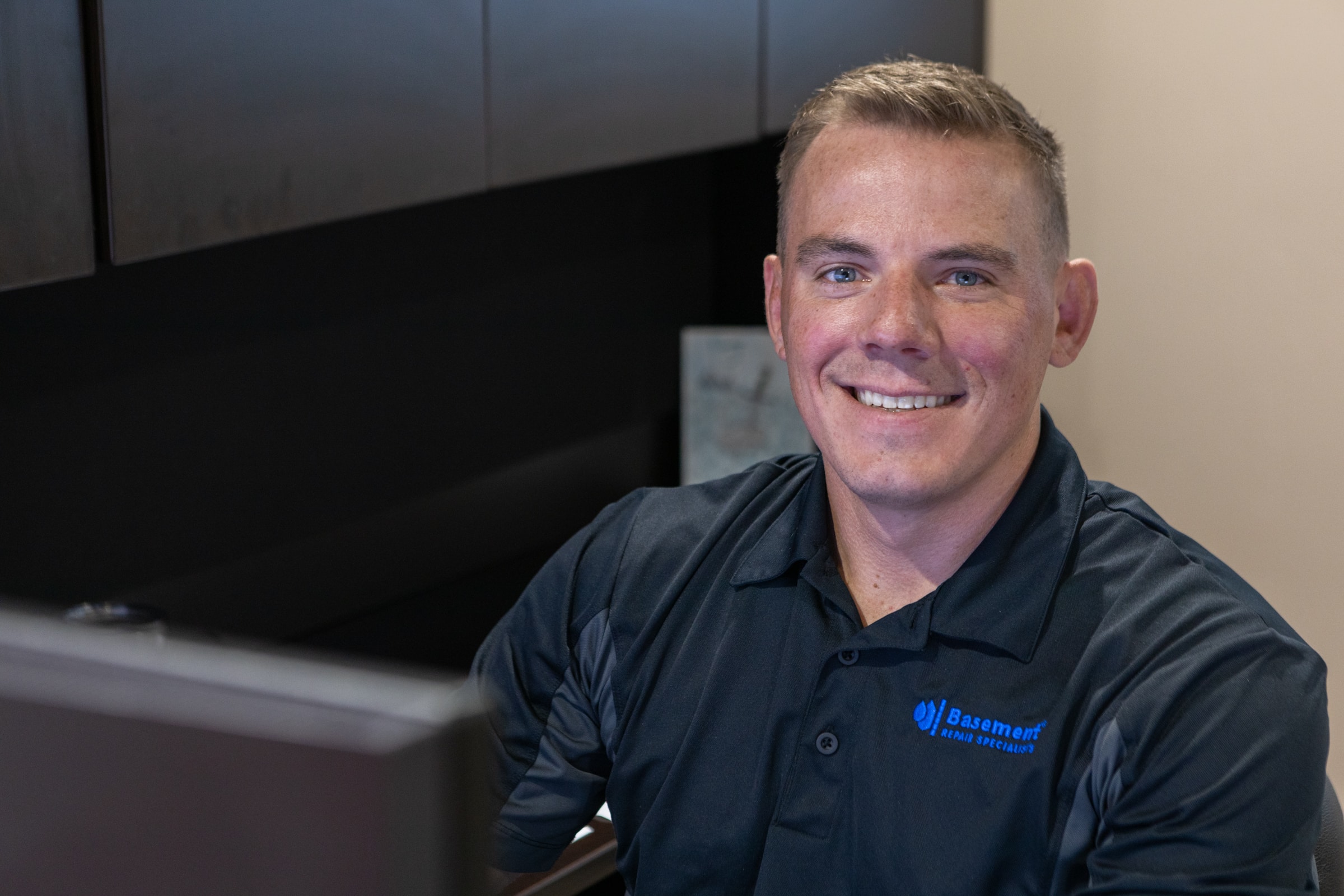 Smiling employee in a Basement Repair Specialists shirt at a desk, showcasing a welcoming work environment and commitment to customer service.