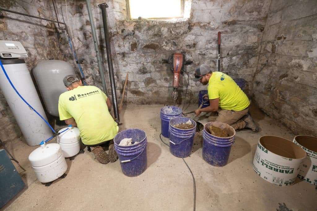 Workers in bright yellow shirts performing basement waterproofing tasks, including mixing materials and using tools, in a stone-walled basement with water management equipment.