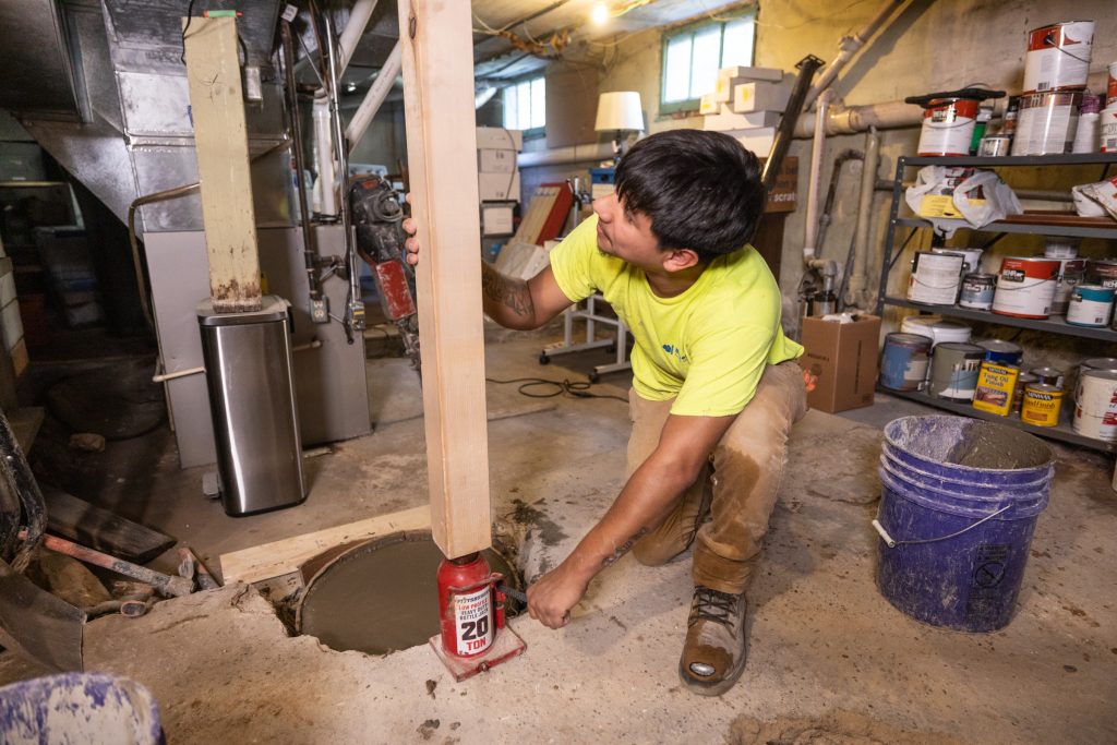 How Basement Waterproofing Prepares You for Resale Worker using a hydraulic jack to install a support column in a basement, surrounded by construction tools and materials, emphasizing basement repair and waterproofing services.