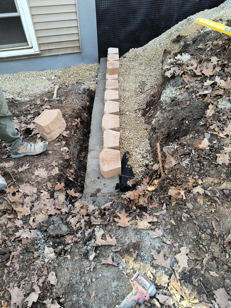 Excavated area for basement waterproofing showing a row of bricks, gravel, and tools, illustrating preparation for drainage solutions in a residential setting.