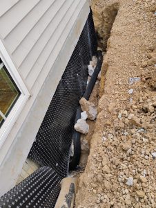 Waterproofing installation featuring a black drainage membrane and gravel surrounding the foundation of a home, illustrating essential moisture barriers for storm protection.