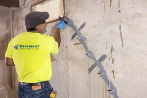 Professional applying crack repair techniques on a basement wall, wearing a bright yellow shirt with "Basement Repair Specialists" branding, emphasizing foundation maintenance and repair.