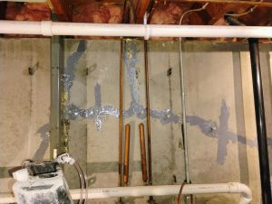 Concrete wall with exposed pipes features patched areas sealed with dark blue material. PVC pipes and insulation visible, suggesting repair work in a basement.