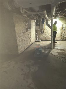 Dehumidifier in basement with stone walls and worker inspecting moisture issues, relevant to basement repair and odor solutions.