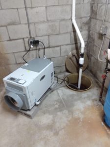 Dehumidifier positioned in a basement corner, connected to a sump pump, addressing moisture issues related to basement odors and waterproofing solutions.