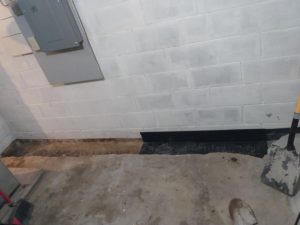 Concrete basement with a white-painted cinder block wall, utility panel, and newly excavated trench along the floor. A shovel leans against the wall.