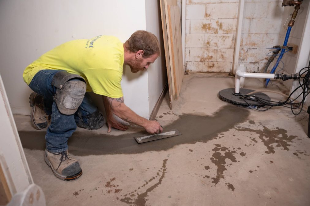 Man applying concrete to the basement floor, preparing for sump pump installation and water management system, with visible sump pump and plumbing in the background.